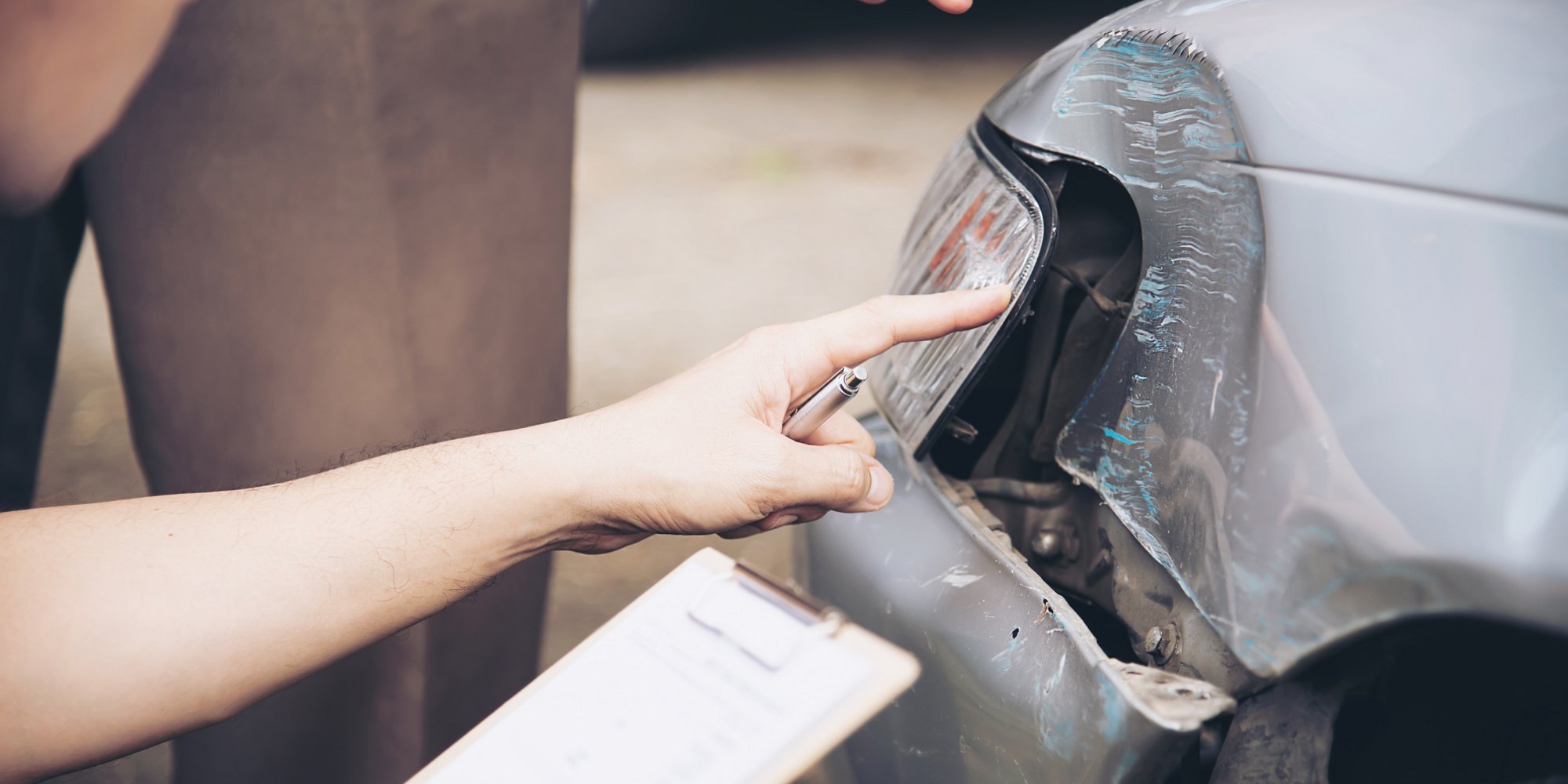 Zwei Personen begutachten den beschädigten Bereich an der Vorderseite eines Autos. Eine Person zeigt mit dem Finger auf den Schaden und hält ein Klemmbrett mit Notizen.