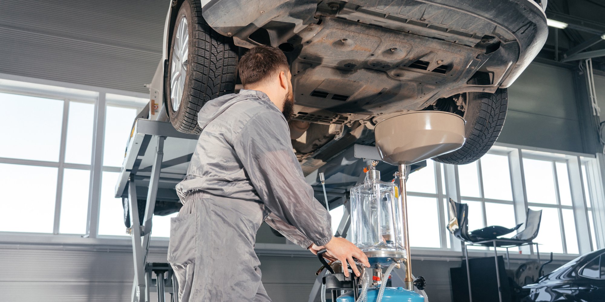 Mechaniker in grauem Overall führt einen Ölwechsel an einem Fahrzeug durch, das auf einer Hebebühne in einer hellen Werkstatt steht.